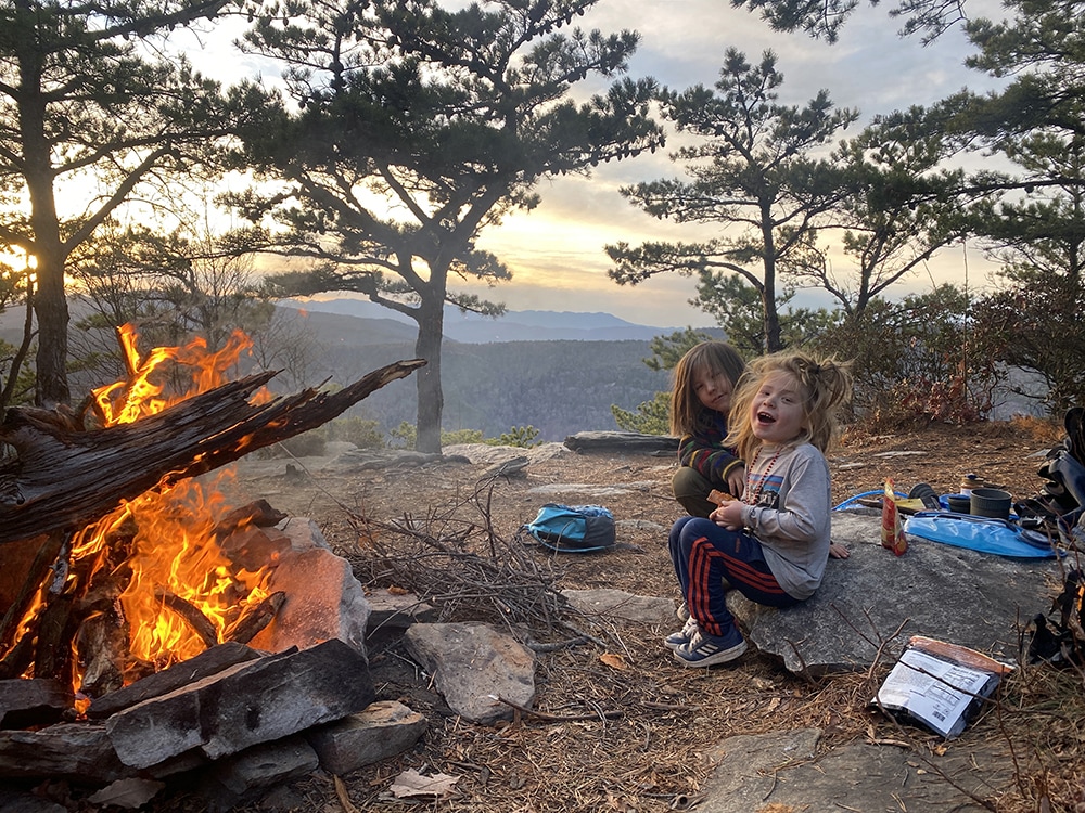 Two children by a campfire at dusk in Linville Gorge, with rolling mountains in the background